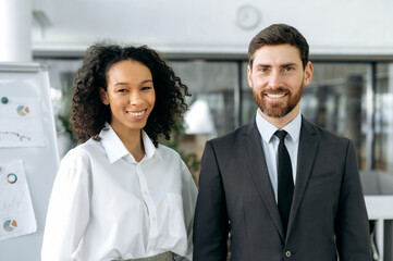 Portrait of two successful multiracial colleagues, company co-founders, directors, managers, a caucasian man and an African American woman, standing in a modern office, looking at camera smiling