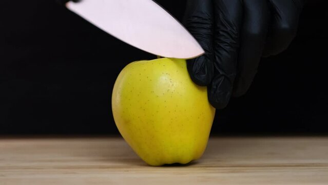 One  yellow apple lies on cutting board. Large kitchen knife cuts a apple into two pieces. Cuts an apple in half with a knife on black background. Human hands in black gloves cut fruits, Close-up slow