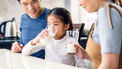 Fototapeta premium Portrait of enjoy happy love asian family father and mother with little asian girl smiling and having protein breakfast drinking and hold glasses of milk at table in kitchen