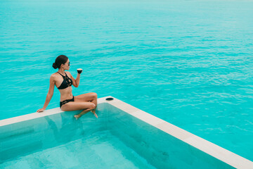 Woman drinking coffee at luxury hotel pool with view of turquoise ocean and overwater infinity...