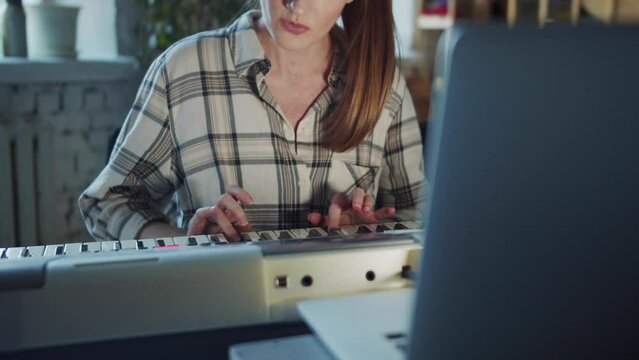 A woman presses the piano keys. She plays with concentration and sometimes looks at the laptop screen