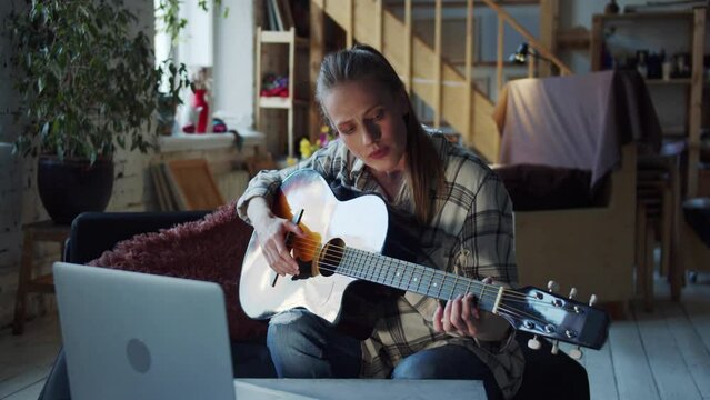 A woman tries to take chords on the guitar and play notes. She is watching videos on her laptop