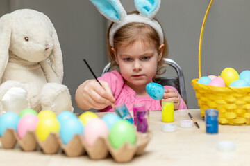 Easter holiday craft at home. Little girl in Easter bunny ears painting colored eggs.