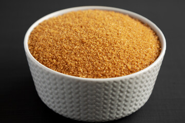 Brown Demerara Sugar in a Bowl on a black surface, side view.