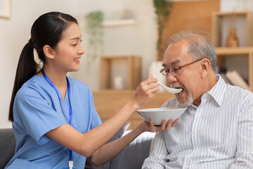 Obraz premium Young Asian nurse feeding breakfast to senior man at couch. Old Asian man with white beard enjoy his meal. Caregiver take care of senior man, give soft food, look after to patient.Elderly Care Concept