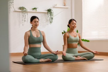 Fotobehang Calm of wellness Couple Asian young woman sit on yoga mat doing breathing exercise yoga lotus pose together.Yoga meditation of two healthy female relax and comfortable at cozy home.Healthy lifestyle © 220 Selfmade studio