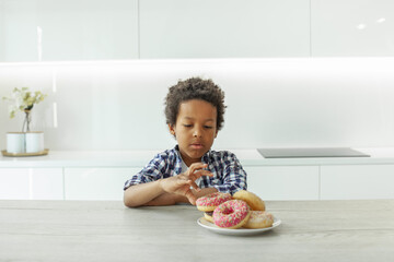 Cute kid boy with black hair eating donuts in white kitchen