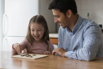 Happy preschool daughter kid and handsome dad playing board game at kitchen table, enjoying home leisure, playtime together. Father teaching cute girl to play checker draughts. Fatherhood concept