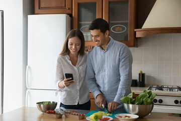 Happy young married couple cooking in home kitchen, checking recipe on mobile phone, ordering food ingredients on internet store. Husband and wife preparing fresh salad for dinner, cutting vegs