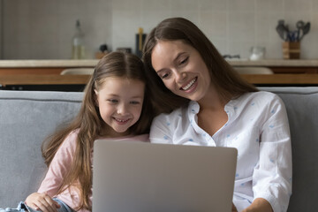 Happy mother and little daughter girl using virtual application on laptop, holding computer, sitting together on couch, reading book, making video conference call, smiling, laughing