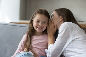 Happy mom whispering in girls ear, sharing secret with joyful kid. Young mother telling funny story to child, enjoying friendship, trust, family leisure, motherhood, having fun with little daughter