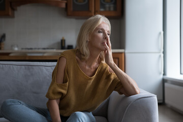 Frustrated lonely mature 50s woman looking away, sitting on sofa at home touching face. Sad lady thinking over health problems, suffering from apathy, depress disorder, memory loss, dementia