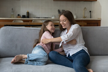 Obraz premium Happy playful mom tickling excited daughter girl on sofa at home, enjoying active games, motherhood. Nanny playing with child on couch, having fun, laughing, giggling. Family, upbringing concept