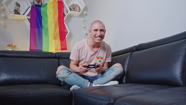 Proud queer caucasian activist using gamepad and winning the game. Rainbow flag in the background. High quality 4k footage