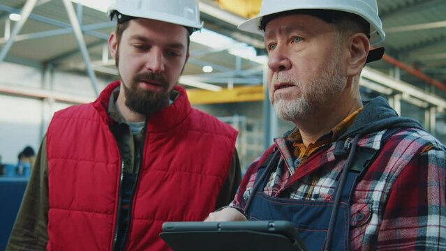 Close up young and elder engineers men heavy industrial workers with helmet using a digital tablet in factory. Checks production processes. Inspector in warehouse.