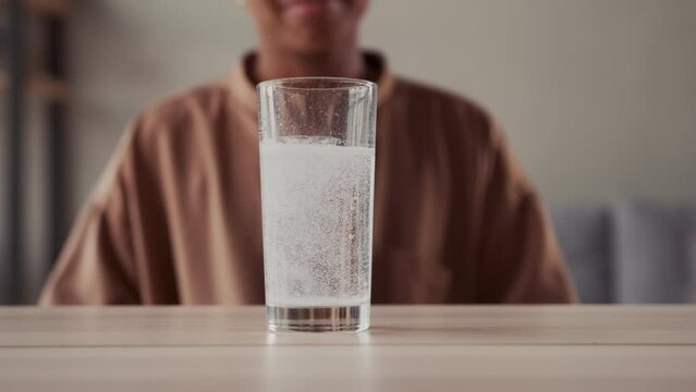 African American woman sits behind glass of water with gas from dissolution of effervescent powder or tablet to get rid of effects of alcohol or health and immune problems which is on table in room