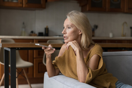 Serious senior woman having mobile phone call on speaker, recording audio message for online chat on smartphone sitting on sofa at home. Mature using voice recognition app on cell