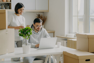 Happy woman and man spend morning at their new kitchen with big windows and modern furniture, husband works on laptop computer discusses with wife trendy design