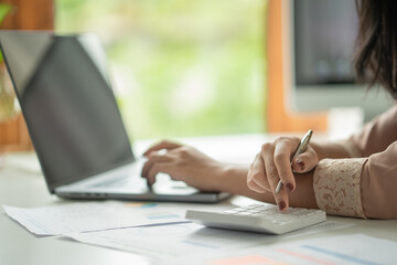 Hand with pen to use calculator and laptop keyboard, graphs and file folders on the desk, business...