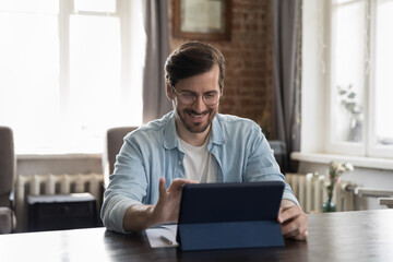 Happy millennial businessman in casual using tablet computer, working in stylish loft office, touching screen, smiling. Freelance worker, employee making video call to client, using virtual app