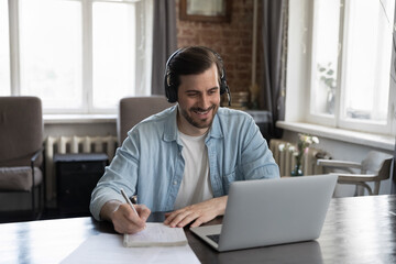 Happy positive adult student in earphones with mic attending virtual class, learning business conference, writing notes, watching webinar on Internet, studying online, making video call