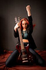 Fototapeta na wymiar Sexy and brutal rocker punk or metal red-hair girl with an electric guitar in a black jacket and jeans is sitting on the floor on her knees in a defiant pose and hand up in the air
