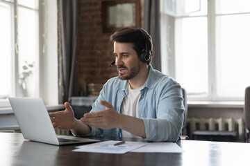 Busy handsome business freelance professional man in headphones talking to client on video call at paper graphic reports on work table. Employee, manager attending internet conference
