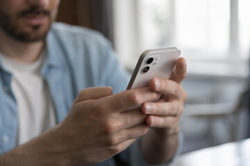 Millennial Caucasian man sitting at table, holding cellphone, typing online chat message on mobile phone. Close up of hands. Guy, business man, office employee making call, browsing on social network