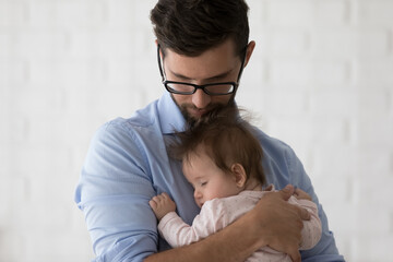 Peaceful young dad in glasses holding sleepy baby daughter in arms, rocking, calming child, enjoying parenthood, fatherhood, childcare, family leisure time at home. Being father concept