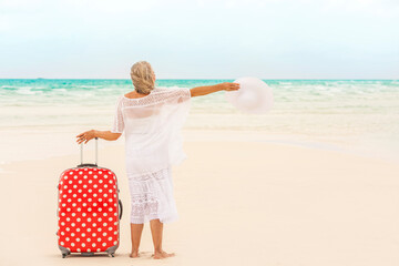 Senior Caucasian female in white on Caribbean beach with red polka dot suitcase