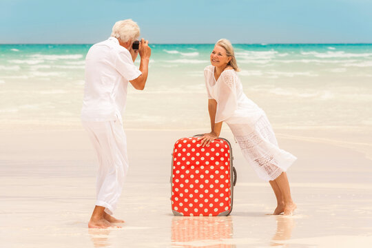 Laughing Caucasian seniors on a tropical beach posing with a camera and suitcase