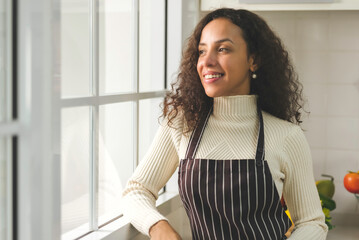 Portrait of beautiful young black woman is standing near window in the bright white kitchen. wife or housewife is preparing to cook healthy food for the family. Stay at home.