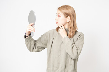 白い背景の前に立って鏡で顔を見る若い女性