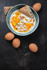 Cilbir or a turkish dish of poached eggs with yogurt, flat lay on a dark-bron stone background, vertical shot with space