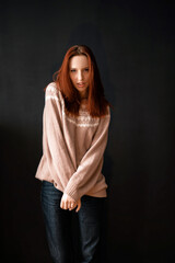 A beautiful slender model girl with fiery red hair, in modest clothes (a warm knitted wrinkled oversize sweater and blue jeans) poses in front of the camera on a black background