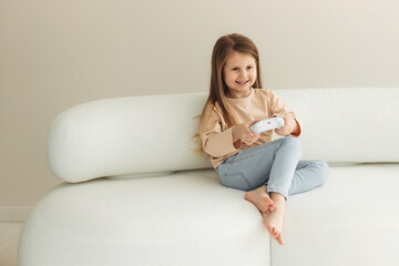 Obraz premium Little smiling girl playing video games while holding a game controller while sitting on a sofa in the living room