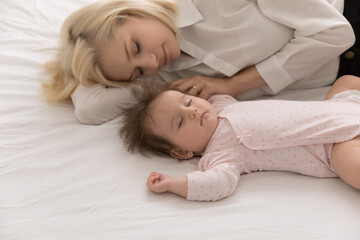 Calm tired new mom and beautiful infant sleeping together, lying in bed on white linen sheet, relaxing in bedroom, taking break, pause. Young mother relaxing near sleepy baby at home. Motherhood