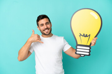 Young handsome caucasian man isolated on blue background holding a bulb icon and doing phone gesture