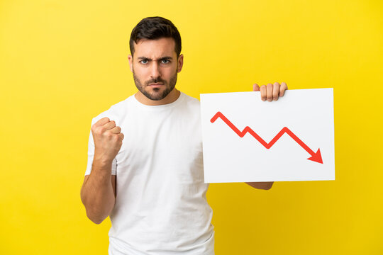 Young handsome caucasian man isolated on yellow background holding a sign with a decreasing statistics arrow symbol and angry