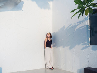 Asian woman look cool with short hair wearing black tank top and white trousers standing and keep hands in pockets on white wall background, full range. Female summer fashion minimal style. © tete_escape