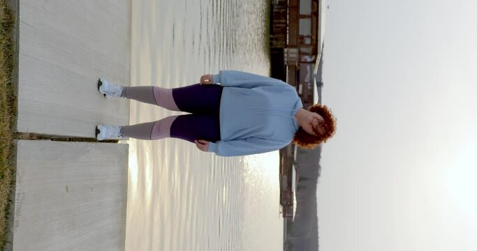 curly girl with red hair in leggings and a blue bike stretches her neck, scrolling her head to the sides, doing an exercise at sunset on the lake. camera rotation. vertical view