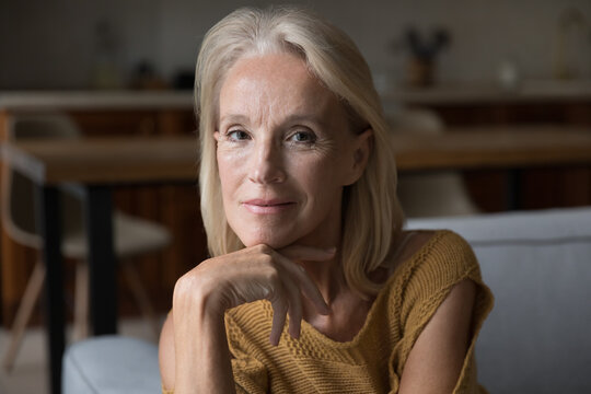 Smiling blonde middle aged 60s lady home head shot portrait. Skinny retired woman, pensioner, OAP sitting on couch, touching chin, looking at camera, posing for profile picture