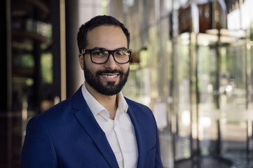 Confident smiling Arabian business man wearing stylish eyeglasses looking at camera standing in modern office. Portrait of handsome Moroccan manager at workplace.  Successful business concept