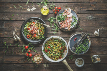Food background with green pasta dish, plates, cooking pan and ingredients on rustic wooden kitchen table. Noodles with arugula, ham, cheese, tomatoes and olive oil. Top view. Italian food