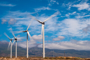 group of windmills lined up on the left with blades in motion. blue sky with clouds.