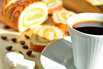 morning home breakfast - cup of strong invigorating black coffee close up on blurred background coffee beans with fresh sweet pastries and sugar cubes on wooden board in sunlight. Selective focus