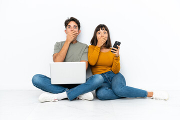 Young couple with a laptop and mobile sitting on the floor covering mouth with hands for saying something inappropriate - obrazy, fototapety, plakaty