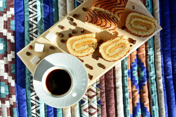 breakfast - cup of strong invigorating black coffee on background of fresh sweet pastries and coffee beans with sugar cubes on wooden board on pattern cloth in bright morning sunlight. Top view