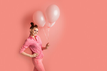 Cheerful redhead woman dressed in pink shirt poses with bunch of inflated balloons laughs out gladully poses on pink background.