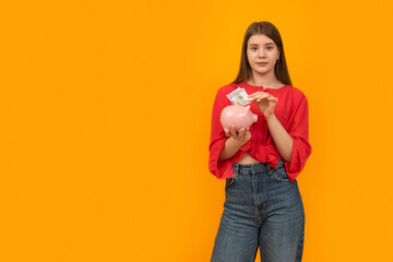 Teenage girl putting dollar bill in piggy bank on yellow background. Financial planning, savings.